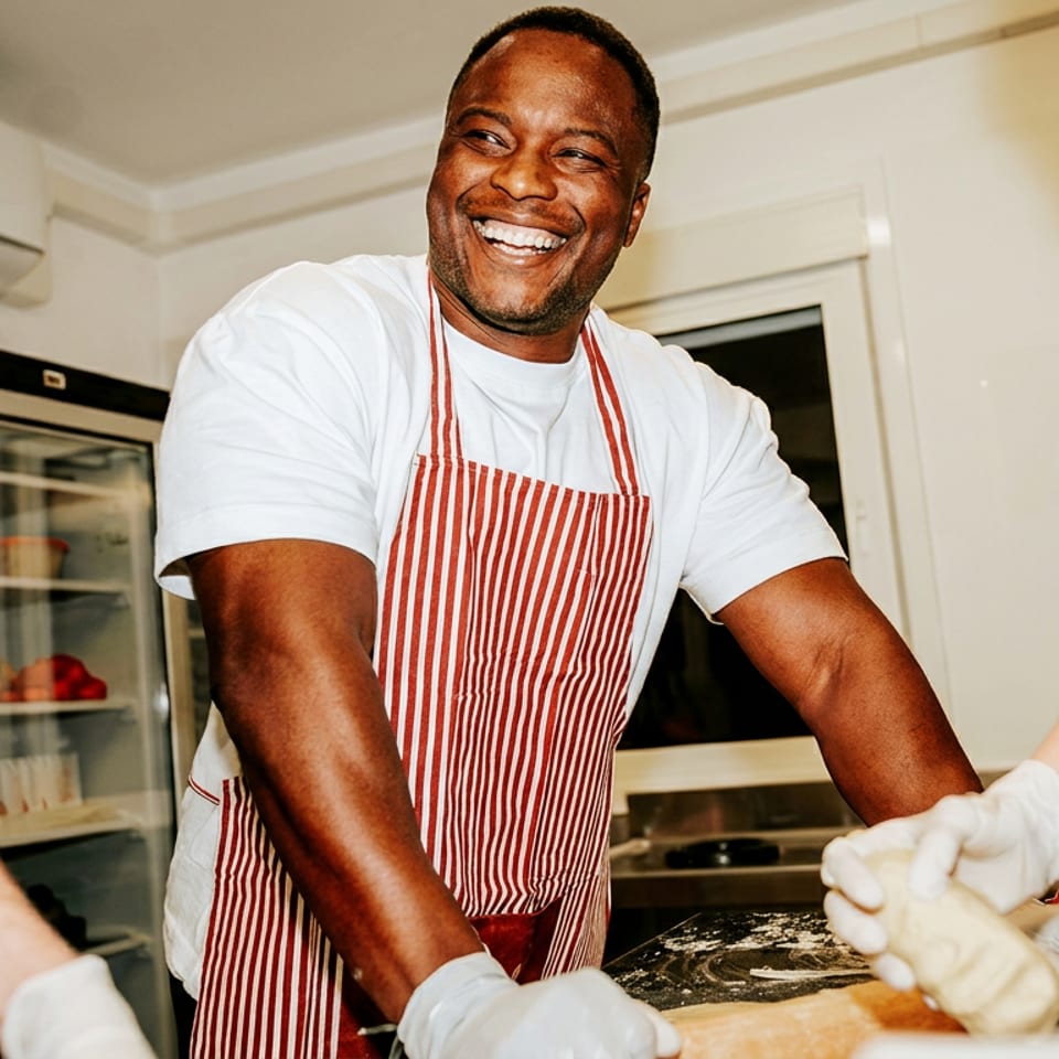 Smiling chef in a red-striped apron prepares dough with gloved teammates in a bright commercial kitchen.